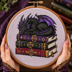 May include: A cross-stitch embroidery in a round wooden hoop featuring a black dragon with purple accents sleeping atop a stack of three books. The books are purple, dark green, and red, each decorated with gold-colored ornate metal clasps.
