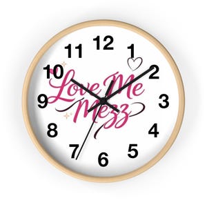 Op de afbeelding: Een ronde wandklok met een lichthouten frame en een witte wijzerplaat. De klok heeft zwarte cijfers van 1 tot 12, zwarte uur-, minuut- en secondewijzers, en de roze cursieve tekst Love Me Mezz met een klein hartje en sterretjes als decoratie.