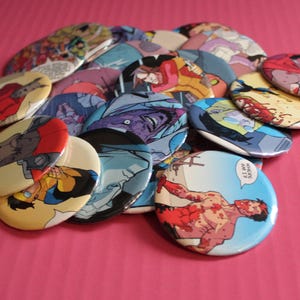 May include: A collection of colorful, oval-shaped buttons featuring comic book art. The buttons display various characters and scenes, with vibrant colors and detailed illustrations. The buttons are scattered on a pink surface.