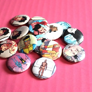 May include: A collection of round buttons with various comic book character illustrations. The buttons feature vibrant colors and detailed artwork, showcasing different characters in action poses. The buttons are scattered on a pink surface.