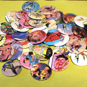 Op de afbeelding: Een verzameling ronde, kleurrijke buttonspins met verschillende stripfiguren. De buttons tonen levendige illustraties van superhelden en andere figuren, met een verscheidenheid aan kleuren en ontwerpen. De buttons liggen op een gele ondergrond.