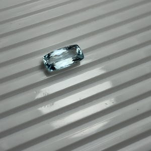 Puede incluir: Una piedra preciosa rectangular facetada con una apariencia translúcida de color azul claro. La piedra presenta bordes afilados y limpios y una superficie pulida que refleja la luz, mostrando su color azul hielo claro y su corte geométrico preciso.