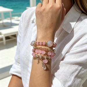 May include: A stack of four beaded bracelets worn on the wrist. The bracelets feature a combination of light pink round beads, translucent faceted beads, gold-toned spacers, and metallic accents. One bracelet includes a dangling clear crystal charm.