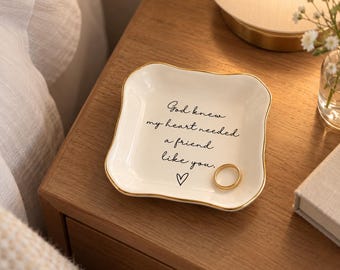 May include: A square white ceramic trinket dish with wavy edges and a gold-painted rim. The center features black cursive text that reads, "God knew my heart needed a friend like you," with a small black heart icon and a gold ring.