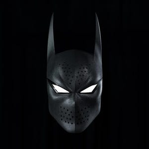 May include: A black full-face mask featuring two tall, pointed ears at the top, almond-shaped eye openings with bright white lenses, and perforated dot patterns across the forehead and nose area, set against a solid black background.