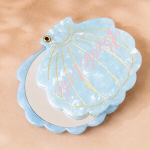 以下が含まれることがあります： パール加工が施された、水色の貝殻型コンパクトミラー。 ミラーはスカラップエッジとゴールドのアクセントが付いています。 「Bride」の文字がピンク色の筆記体でミラー上部に書かれています。