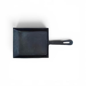Puede incluir: Una pequeña sartén de hierro fundido negro con una superficie de cocción rectangular y un mango corto. La sartén está diseñada para porciones individuales o pequeñas. El mango tiene un agujero para colgar. La sartén está sobre un fondo blanco.