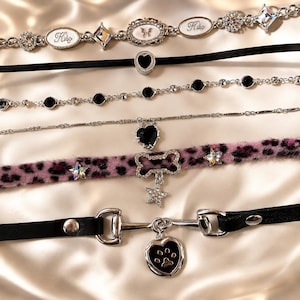 May include: A collection of six necklaces including a silver chain with oval charms, a black velvet choker with a heart, a crystal-studded chain, a thin silver chain with a heart, a fuzzy pink leopard print choker with a bone and star, and a black leather strap with a heart charm.