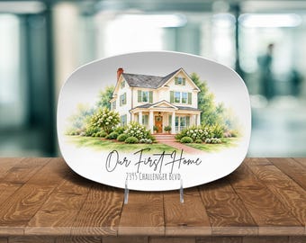 May include: A white oval ceramic plate featuring a watercolor illustration of a two-story house with green shutters and a brick walkway. The text "Our First Home" and "2395 CHALLENGER BLVD." is printed in black script and sans-serif font.