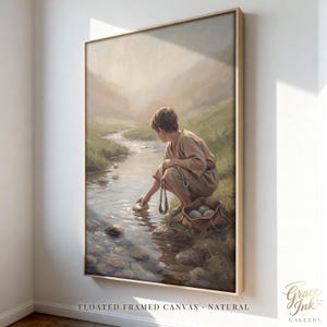 May include: A framed canvas print depicting a person in tan clothing crouching by a stream, reaching into the water. Beside them sits a brown leather pouch filled with stones. The scene features soft, muted tones of green, brown, and beige.