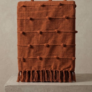 May include: A folded rust-colored throw blanket with a textured weave and knotted details. The blanket features horizontal rows of knotted accents and a fringe trim along the bottom edge. The throw is displayed on a concrete surface.
