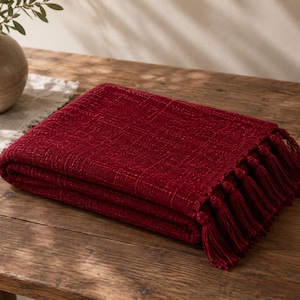 May include: A deep red throw blanket with a textured, woven fabric construction and a series of matching red tassels attached along the edge. The blanket is folded into a rectangular shape, showcasing the intricate weave and the fringe detail.