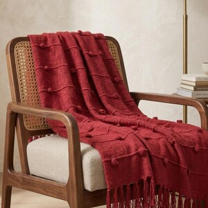 May include: A deep red throw blanket featuring a textured knit pattern with raised pom-pom accents arranged in horizontal rows. The blanket is finished with long, matching red fringe along the bottom edge, creating a decorative and tactile appearance.
