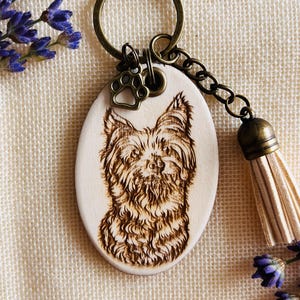 May include: An oval wooden keyring featuring a detailed pyrography portrait of a Yorkshire Terrier. The keyring includes a bronze-toned metal ring, a small paw print charm, and a light beige faux leather tassel attached by a chain.