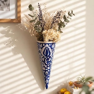May include: A blue and white ceramic wall vase with a floral design, filled with dried flowers and greenery. The cone-shaped vase hangs on a white wall. The vase is approximately 30 cm tall.