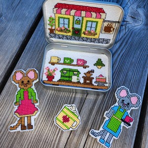 May include: A metal tin containing cross-stitch embroidery of a cafe interior, with two separate mouse-shaped cross-stitch figures and a cross-stitch coffee mug with whipped cream placed outside the open container on a wooden surface.
