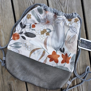 May include: A drawstring backpack featuring a cream-colored upper section with a botanical print of orange, gray, and brown flowers and leaves. The lower portion is made of solid gray material. It includes gray rope straps and a small white tag.