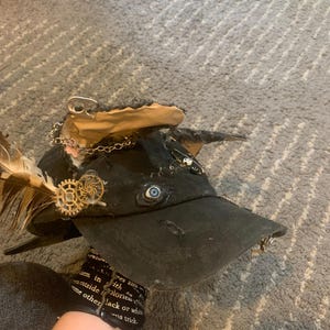 以下が含まれることがあります： ギア型の金属製チャーム、リアルな青い目、銀色のチェーン、缶のプルタブ、茶色の羽など、さまざまな装飾が施された黒い野球帽。キャップの縁はほつれており、使い古されたような質感の生地で作られています。