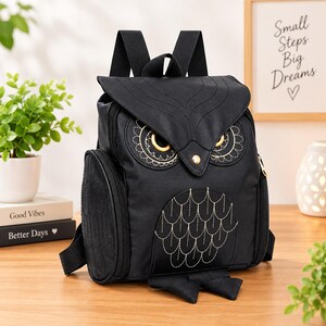 Könnte beinhalten: Ein schwarzer Rucksack in Eulenform mit gestickten goldfarbenen Augendetails und aufgestickten Federmustern auf der Frontklappe und dem Korpus. Der Rucksack verfügt über verstellbare schwarze Schultergurte, Seitentaschen und einen Klappverschluss.