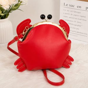 Peut inclure: Un sac bandoulière rouge vif en forme de crabe avec une finition lisse. Le sac présente une armature en métal doré, une fermeture à fermoir avec deux perles sphériques noires et une bandoulière rouge assortie fixée par des attaches dorées.