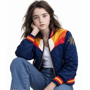 May include: A quilted navy blue bomber jacket featuring a white tank top and blue jeans. The jacket has a gold zipper, ribbed cuffs with red and yellow stripes, and a chevron pattern across the chest in white, orange, and yellow. A gold sun emblem is embroidered on the left sleeve.