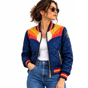 Könnte beinhalten: Eine gesteppte Bomberjacke in Marineblau mit einer sonnenuntergangsartigen Grafik auf der Brust in Gelb, Orange und Pink. Die Jacke hat einen goldfarbenen Reißverschluss, gerippte Bündchen mit roten und gelben Streifen sowie eine kleine Sonnenstickerei.