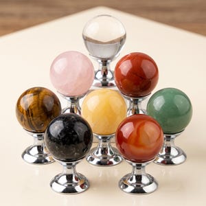 May include: A collection of seven spherical cabinet knobs featuring polished gemstone and clear glass spheres mounted on silver-toned metal bases. The set includes tiger's eye, rose quartz, clear glass, red jasper, yellow jade, black stone, and green aventurine.