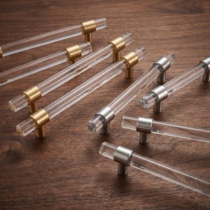 May include: A collection of cabinet pulls featuring clear cylindrical acrylic bars with metallic end caps and mounting posts. The set includes variations with brushed gold-tone and brushed silver-tone metal finishes, arranged in a group.