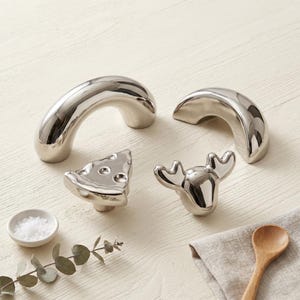 May include: A collection of four silver-toned metal cabinet knobs featuring two smooth arched handles, one knob shaped like a wedge of cheese with circular indentations, and one knob shaped like the head of a moose with prominent antlers.