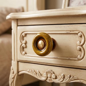 May include: A round, textured, mustard-yellow ceramic drawer knob with a smooth, dark brown inner ring and a central metallic gold-toned circular accent, attached to a cream-colored wooden drawer front with carved decorative scrollwork details.