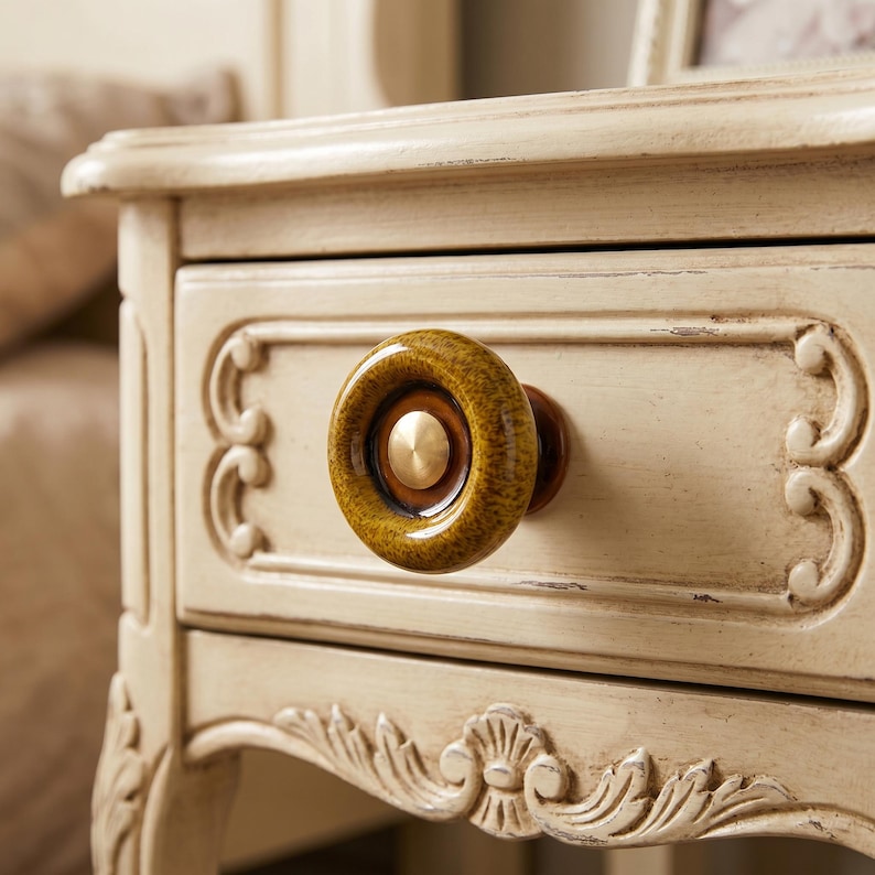 May include: A round, textured, mustard-yellow ceramic drawer knob with a smooth, dark brown inner ring and a central metallic gold-toned circular accent, attached to a cream-colored wooden drawer front with carved decorative scrollwork details.