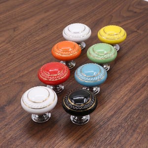 Op de afbeelding: Een verzameling van acht ronde keramische kastknoppen in diverse kleuren, waaronder wit, geel, oranje, groen, rood, blauw en zwart. Elke knop heeft een centraal cirkelvormig detail met een goudkleurige rand en een textuurrijke afwerking.