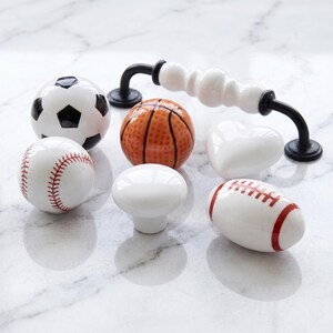 May include: A collection of ceramic cabinet knobs and a drawer pull. The set includes a soccer ball, a basketball, a baseball, a football, a plain white round knob, a white heart-shaped knob, and a white drawer pull with a black metal handle.
