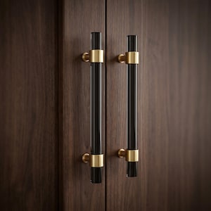May include: Two vertical cabinet pulls featuring cylindrical black glass bars secured by brushed brass metal mounts. The hardware is attached to a dark brown wood surface with a visible vertical grain pattern, creating a contrast between the dark glass and metal.