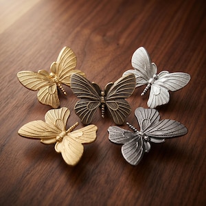 May include: Five butterfly-shaped metal knobs arranged in a cluster. The set includes two gold-toned, one silver-toned, one dark bronze-toned, and one dark grey-toned butterfly, each featuring detailed wing patterns and a central body segment.