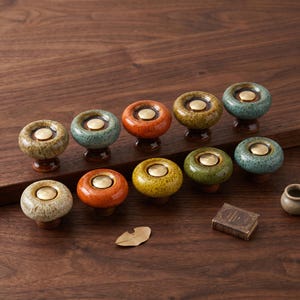May include: A collection of ten round ceramic cabinet knobs featuring a glossy, speckled glaze in various colors including orange, yellow, green, and blue. Each knob has a smooth, circular brass-colored metal insert in the center.