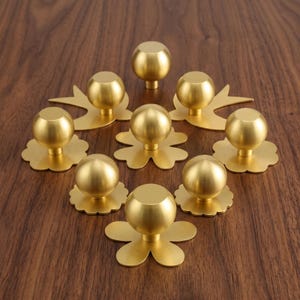 Peut inclure: Un ensemble de neuf boutons de placard en métal doré, chacun doté d'une tête sphérique montée sur une plaque de base décorative plate. Les plaques varient en forme, incluant des motifs de fleurs et de silhouettes d'oiseaux, avec une finition mate.