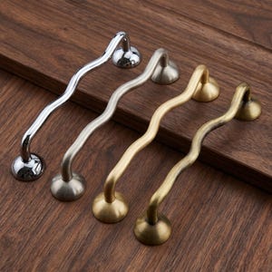May include: Four wavy cabinet pulls arranged in a row, featuring distinct metallic finishes including polished chrome, brushed nickel, matte gold, and an antique brass patina, each with two circular mounting bases at the ends.