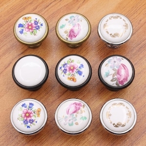 May include: A collection of nine round cabinet knobs featuring white ceramic centers with various floral patterns in pink, blue, and yellow, or gold filigree designs, set within brass, black, or silver-toned metal circular frames and bases.