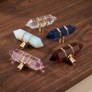 Könnte beinhalten: Fünf doppelendige, kristallförmige Schrankknöpfe in Transparent, opakem Hellblau, Dunkelblau, Rosa und Braun. Jeder Knopf verfügt über einen goldfarbenen Metallsockel und zwei goldfarbene Drahtwicklungen um den kristallartigen Körper.