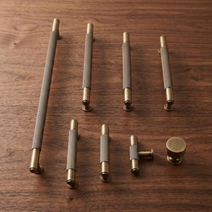 Può includere: Una collezione di maniglie in ottone per mobili che include barre cilindriche di varie lunghezze con una texture a coste verticali, una maniglia a T e un pomello rotondo, tutti caratterizzati da una finitura metallica spazzolata.
