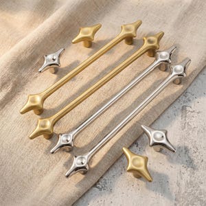 Könnte beinhalten: Eine Sammlung von Möbelbeschlägen, bestehend aus zwei langen goldfarbenen Metallgriffen, zwei langen silberfarbenen Metallgriffen und vier passenden sternförmigen Knöpfen in Gold- und Silberoptik, alle mit einer glatten, polierten Metalloberfläche.