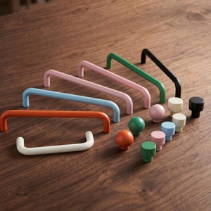 Puede incluir: Una colección de herrajes para muebles que incluye varios tiradores de cajón en forma de U y pomos redondos en varios colores sólidos como naranja, azul claro, rosa, verde, negro y crema, dispuestos en un grupo sobre una superficie.
