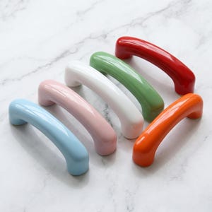 Può includere: Un set di cinque maniglie per armadietti arcuate con una finitura liscia e lucida. Le maniglie sono disposte in una fila diagonale e presentano colori solidi distinti: azzurro, rosa tenue, bianco, verde salvia, rosso acceso e arancione.
