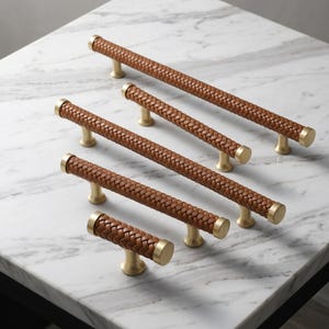 May include: A collection of four cabinet pulls in varying lengths, each featuring a brown braided leather handle wrapped around a central rod, finished with brushed gold metal end caps and mounting posts for attachment to furniture doors.
