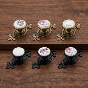 May include: Six drawer knobs with ornate metal backplates. The top row features three knobs with antique brass finish and floral or gold-rimmed white ceramic tops. The bottom row features three knobs with matte black finish and floral or plain white tops.