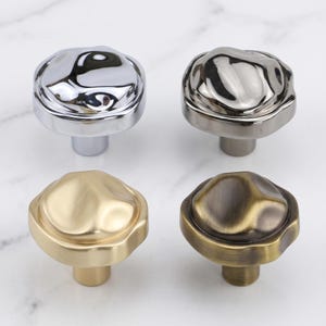May include: Four cabinet knobs with irregular, organic, rounded shapes. The top left knob is polished chrome, the top right is dark gunmetal, the bottom left is brushed gold, and the bottom right is an antique brass finish with dark highlights.
