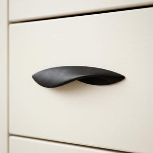 May include: A matte black, curved drawer pull handle mounted on a smooth, off-white wooden drawer front. The handle features a sleek, elongated, and tapered design that extends horizontally across the center of the drawer panel.