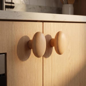 Op de afbeelding: Twee ovaalvormige kastknoppen van licht hout bevestigd op lichtgekleurde houten kastdeuren. De knoppen hebben een glad, afgerond oppervlak en een natuurlijke houtnerfafwerking, horizontaal geplaatst in het midden van elk kastdeurpaneel.