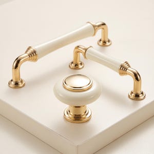 May include: Two gold and cream cabinet pulls and one matching round cabinet knob. The pulls feature a cylindrical cream centre with gold metal ends and ribbed detailing. The round knob has a cream base with a polished gold circular centre.